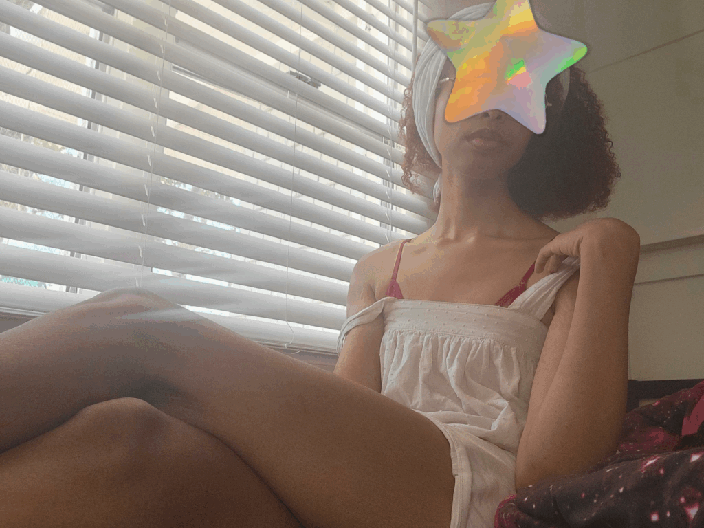Camara Noelle, a brown skinned woman with a star over her face and her longs legs crossed in front of the camera. she's wearing a red lingerie top and a white flared camisole top. she's posing for her photo. this image is for msnovasweets' escorting and camgirl page